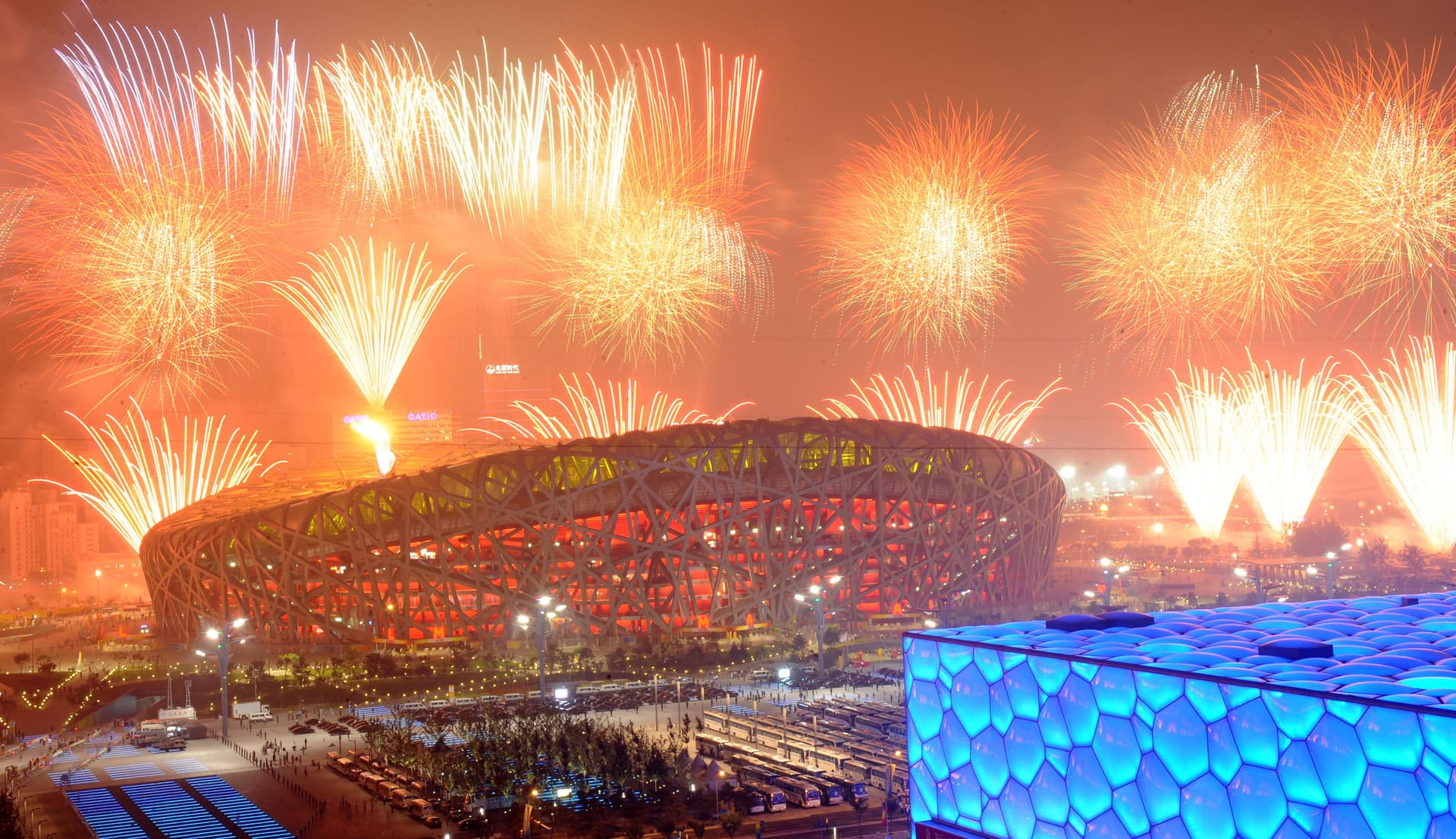 Image is a photograph of the Beijing Olympics stadium during the opening ceremony. Fireworks in the sky and large crowds are visible.